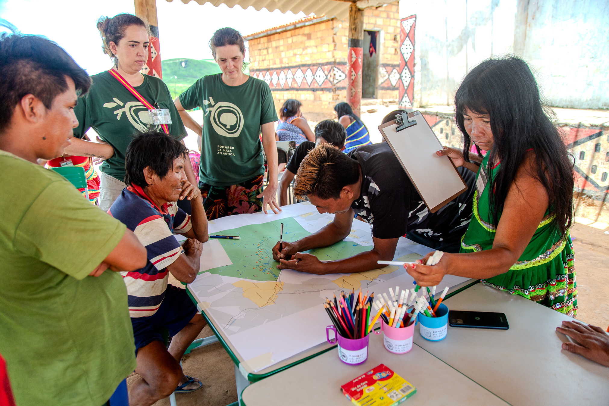 Four Indigenous community members (three men and a women) focus on a map of the region; one of them writes on the map, while two Arboretum Program officers oversee the activity.