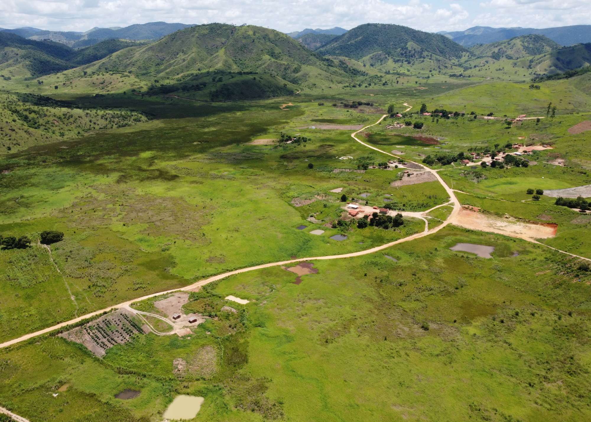 An aerial view of a rural landscape with rolling green hills, scattered trees, a dirt road winding through the terrain, and a small village with red-roofed buildings.